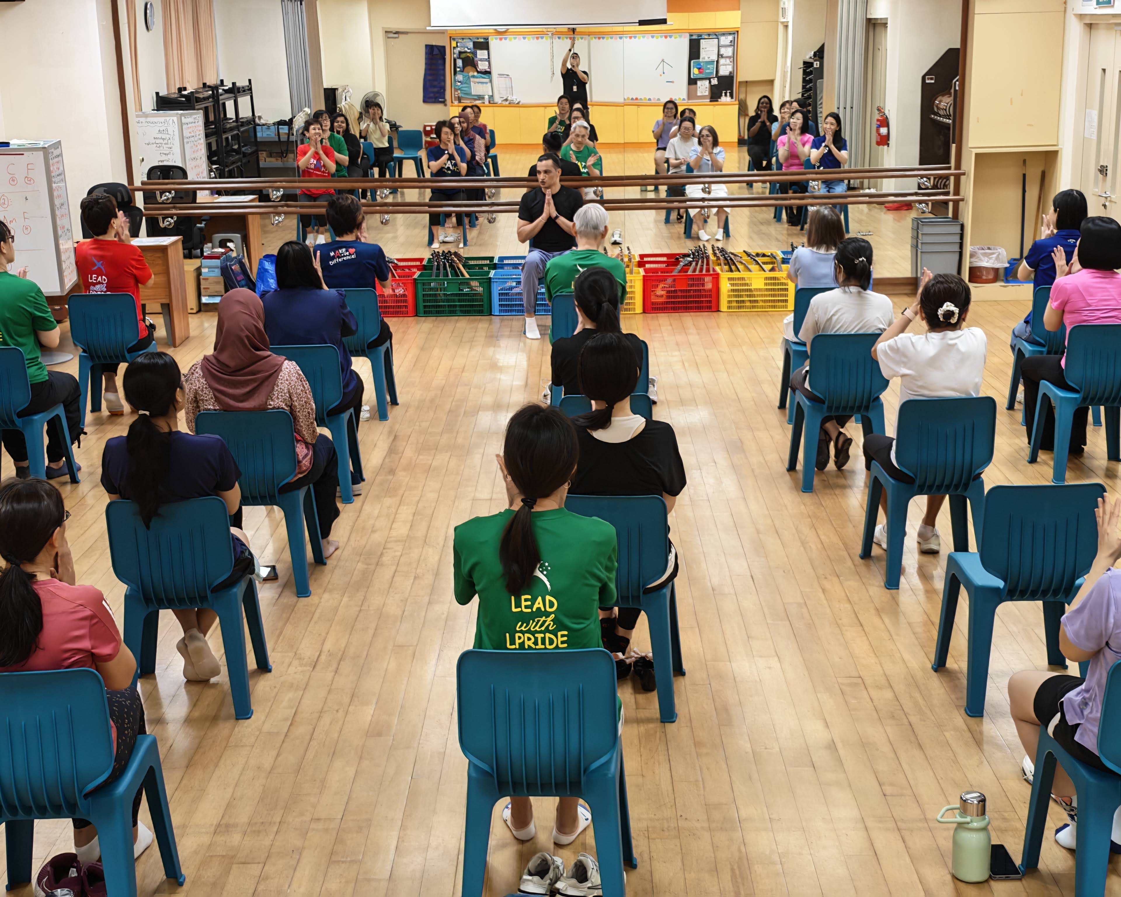 A large group of staff seated on chairs in a dance room follow a yoga instructor standing at the front of the room. The instructor has his hands pressed together in a prayer pose as participants follow along.